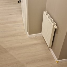 KG Koncept Kacper Gucz - Jasne panele podłogowe po renowacji w Zamościu, widok z góry. Ściany pomalowane na szaro, białe listwy przypodłogowe i grzejnik. Częściowo widoczna biała stolarka drzwiowa.