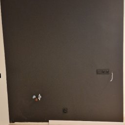 KG Koncept Kacper Gucz - Ciemna ściana po malowaniu z widocznymi gniazdkami, rurami i listwą przypodłogową. Na podłodze jasne panele. Fragment folii ochronnej na górze ściany.