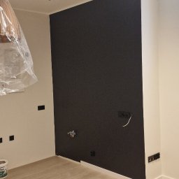 KG Koncept Kacper Gucz - Pokój po remoncie: czarna ściana kontrastuje z jasnymi panelami podłogowymi i białymi ścianami. Widoczne gniazdka elektryczne i lampy sufitowe. Część lampy zakryta folią.