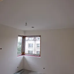 Wnętrze pokoju po położeniu gładzi, widoczne ślady po puszkach elektrycznych, okno narożne z widokiem na budynek mieszkalny.