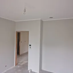 Wnętrze pokoju w szarych odcieniach z widocznymi śladami po remoncie, surowy beton na podłodze, widoczne kable od oświetlenia, listwa przypodłogowa przy suficie.