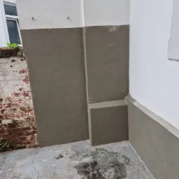 Narożnik zewnętrzny budynku z fragmentem ściany z cegły po lewej, dolna część ściany pokryta szarą warstwą, górna białą, na podłożu widoczne plamy.