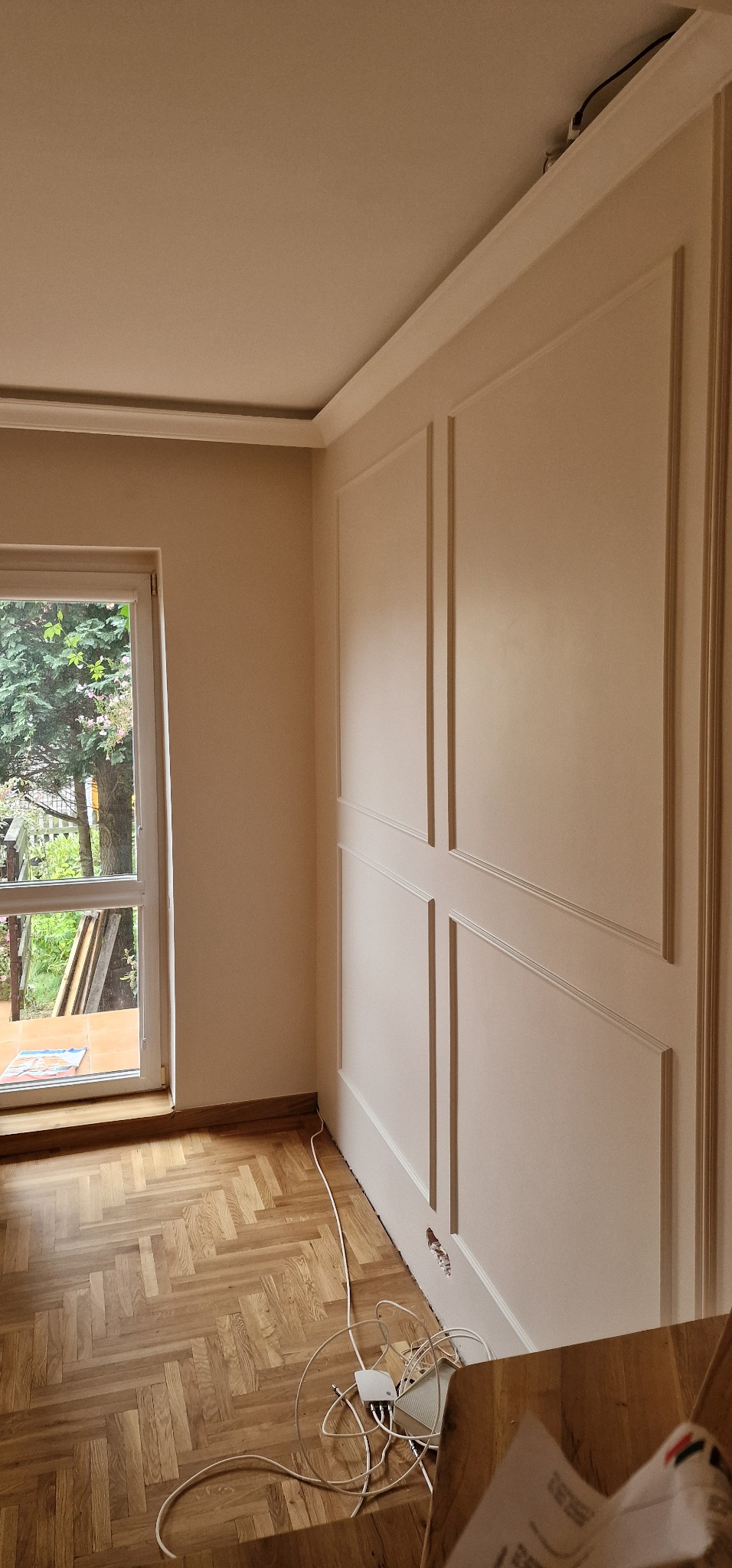 Jasne wnętrze po remoncie: parkiet, okno z widokiem na zieleń i ściana z dekoracyjnymi panelami w stylu angielskim. Widoczne gniazdko elektryczne i przewody.