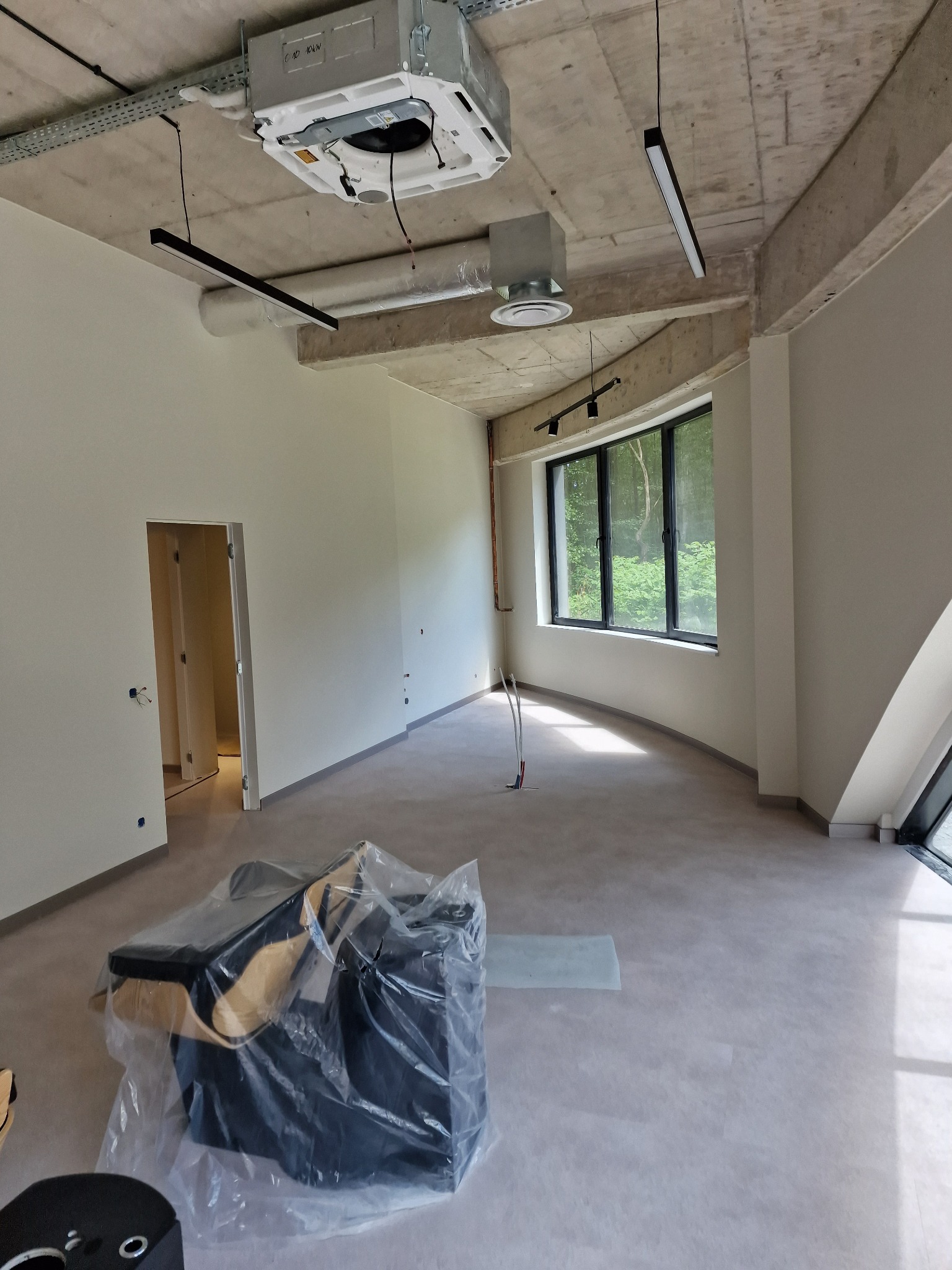 Jasne, przestronne wnętrze biurowe z surowym betonowym sufitem, nowoczesnym oświetleniem i dużymi oknami. Ściany pomalowane na jasny kolor, podłoga szara. Częściowo zakryte meble.