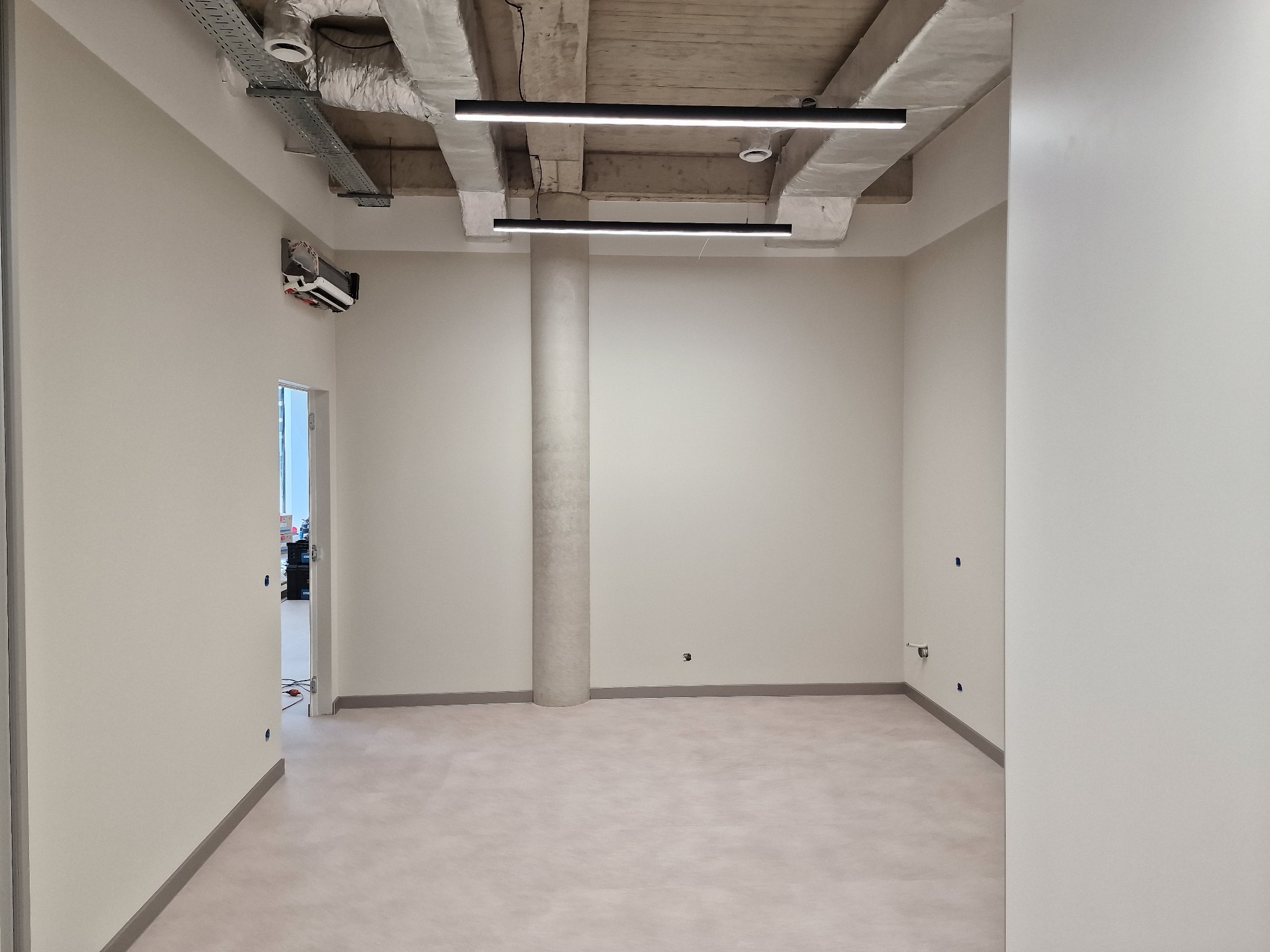 Jasne, przestronne wnętrze biurowe z kolumną, surowym betonowym sufitem z oświetleniem liniowym i klimatyzacją. Widoczne ślady po pracach wykończeniowych na ścianach.