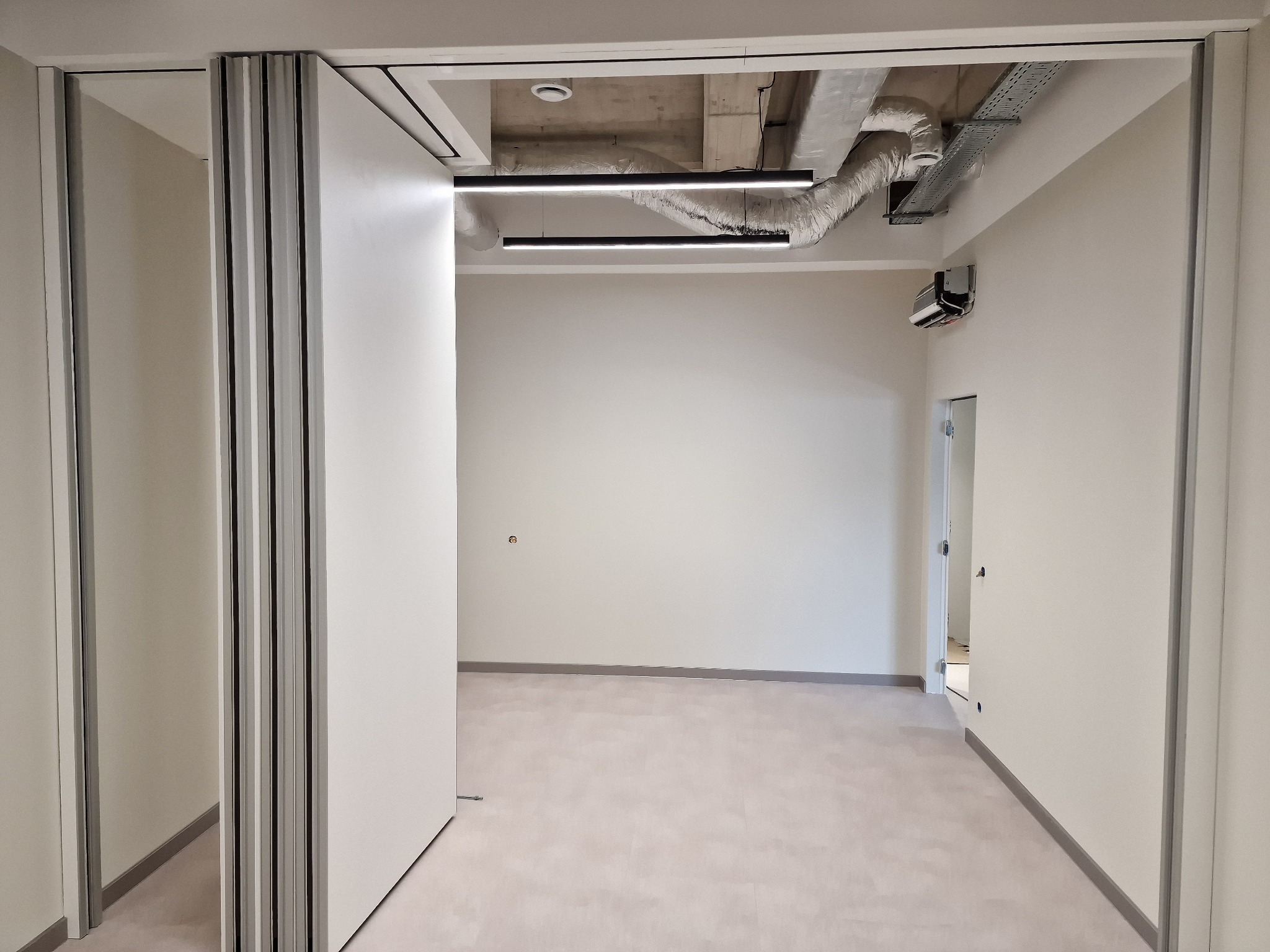 Białe, przesuwne drzwi harmonijkowe w jasnym, minimalistycznym wnętrzu biurowym z widocznymi instalacjami pod sufitem i szarą podłogą.