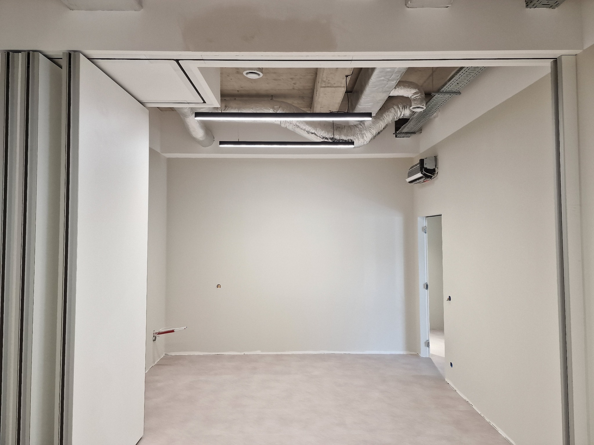 Wnętrze po remoncie: gładkie, jasne ściany, betonowy sufit z instalacjami, nowoczesne oświetlenie liniowe. Widoczne drzwi harmonijkowe i fragment sąsiedniego pomieszczenia.