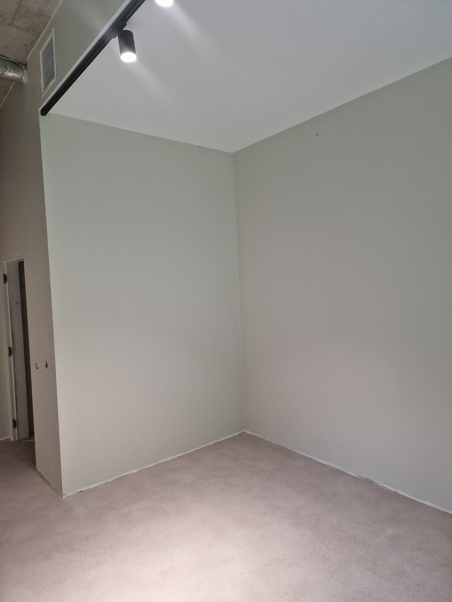 Pomalowane na szaro wnętrze pokoju z zamontowanym oświetleniem szynowym i widoczną podłogą. Świeżo wykończone ściany i podłoga sugerują remont.