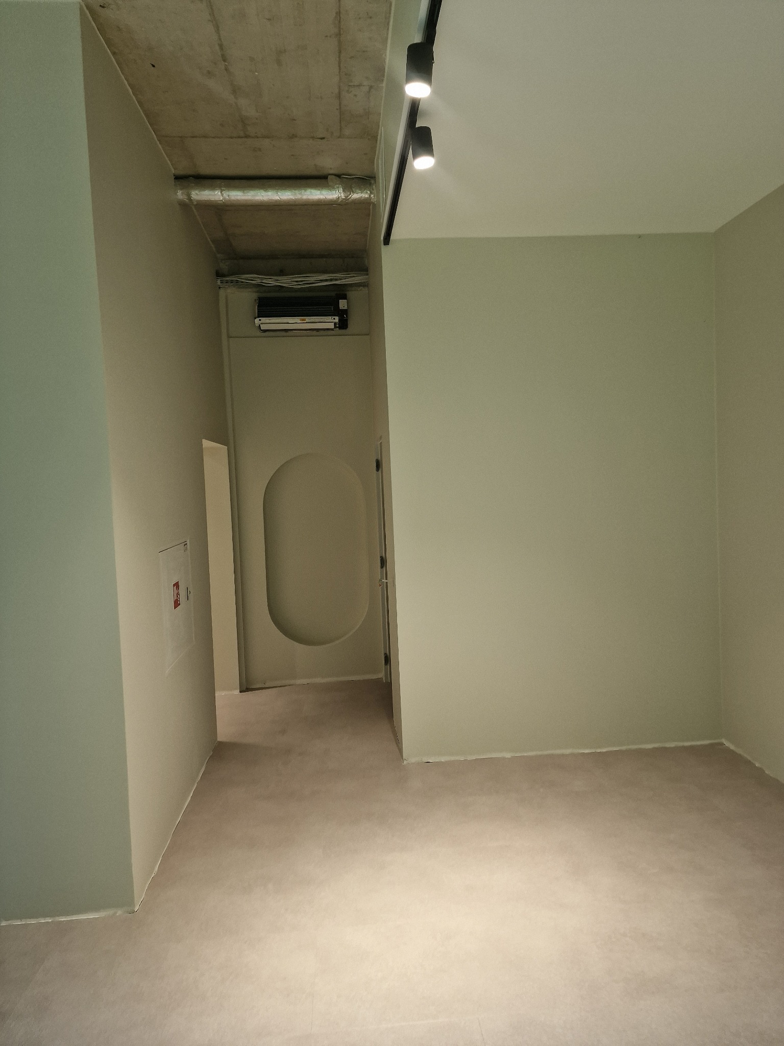 Pomalowane na jasno pomieszczenie z betonowym sufitem i oświetleniem szynowym. Widoczne drzwi z owalnym przeszkleniem i fragment instalacji wentylacyjnej.