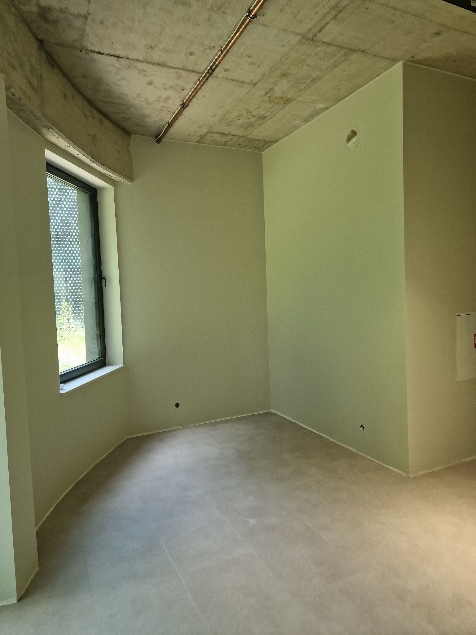 Pomalowane na jasnozielono wnętrze z surowym betonowym sufitem i miedzianymi rurami. Okno z widokiem na zieleń. Podłoga wyłożona jasnymi płytami. Minimalistyczny design.