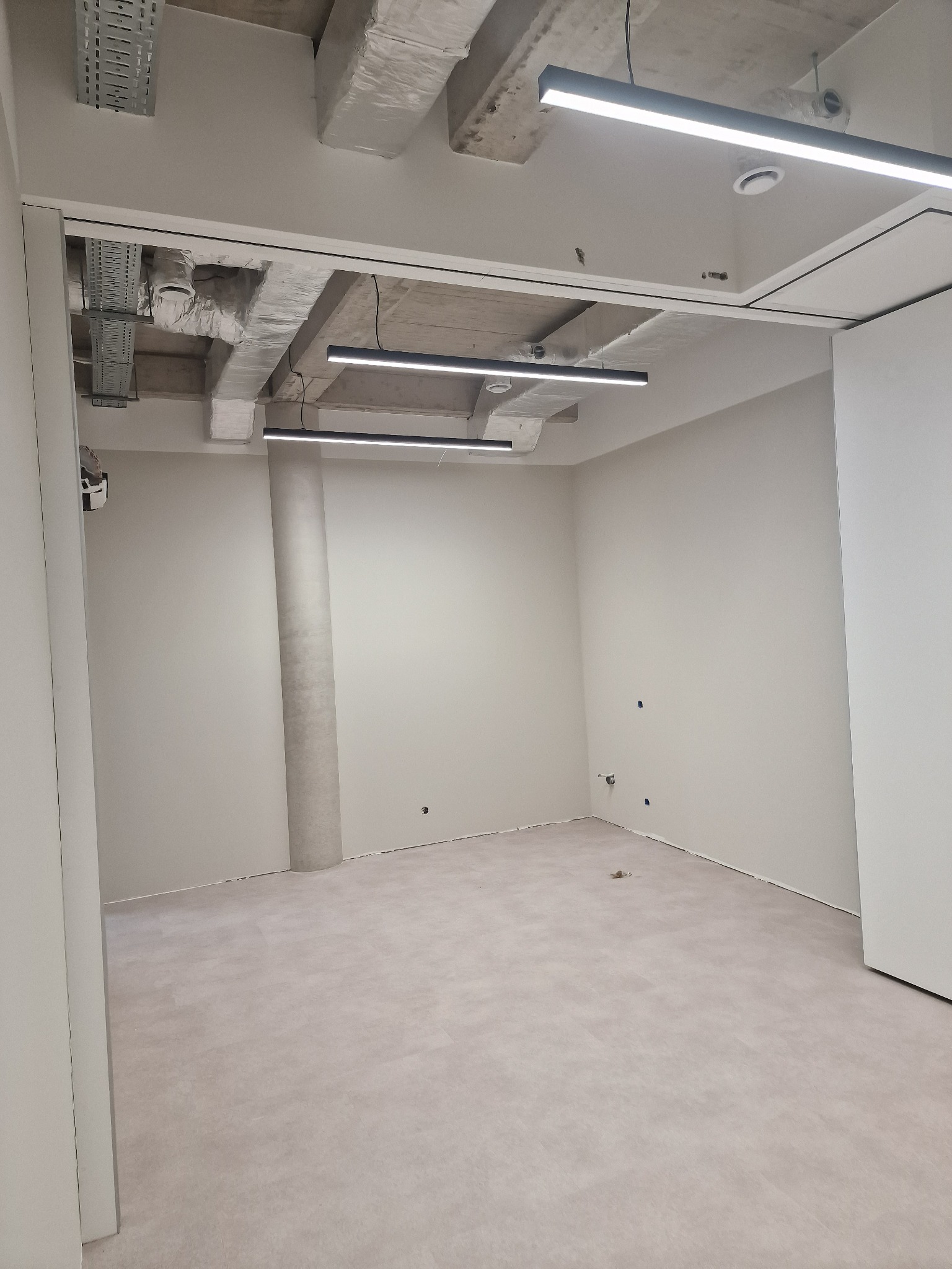 Jasne, puste pomieszczenie biurowe po remoncie, z widocznymi elementami instalacji pod sufitem i kolumną. Ściany pomalowane na jasny odcień, podłoga wyłożona panelami.