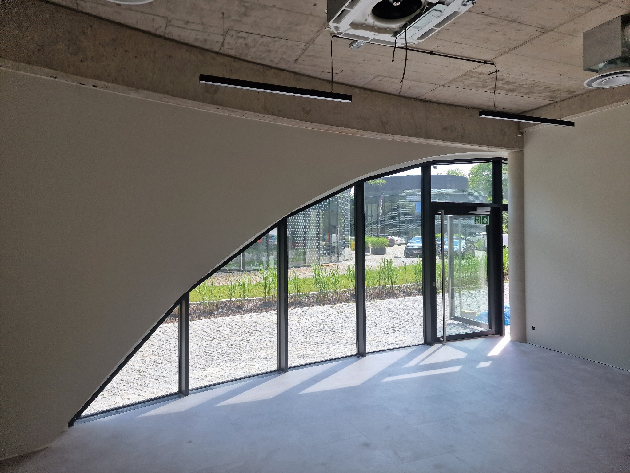 Wnętrze biura po remoncie: gładkie ściany w odcieniach szarości, duże okno z widokiem na zewnątrz, surowy betonowy sufit z nowoczesnym oświetleniem.