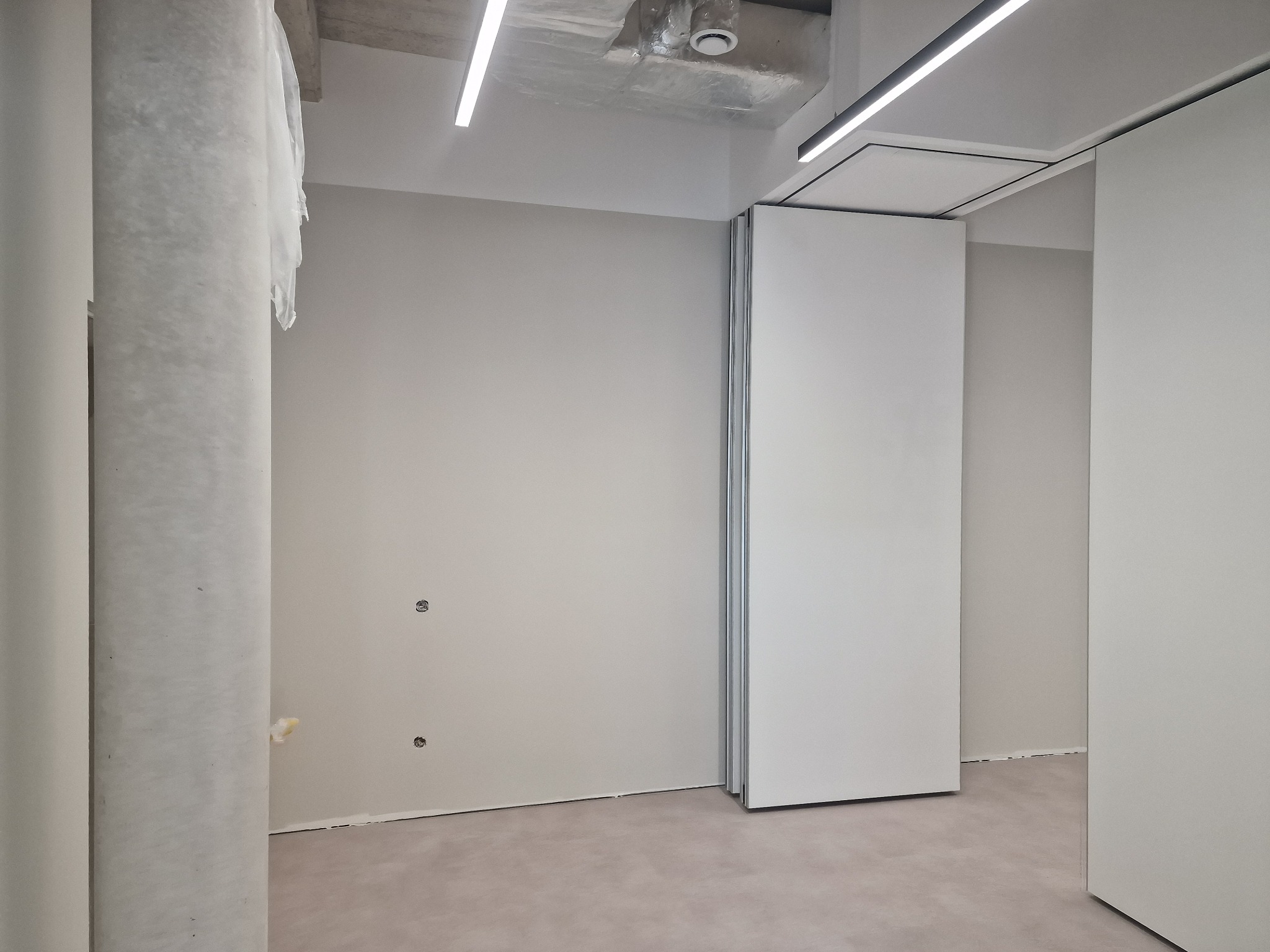 Wnętrze biura po szpachlowaniu i malowaniu ścian na szaro, widoczne przygotowania do montażu drzwi przesuwnych. Surowy betonowy sufit z oświetleniem liniowym.