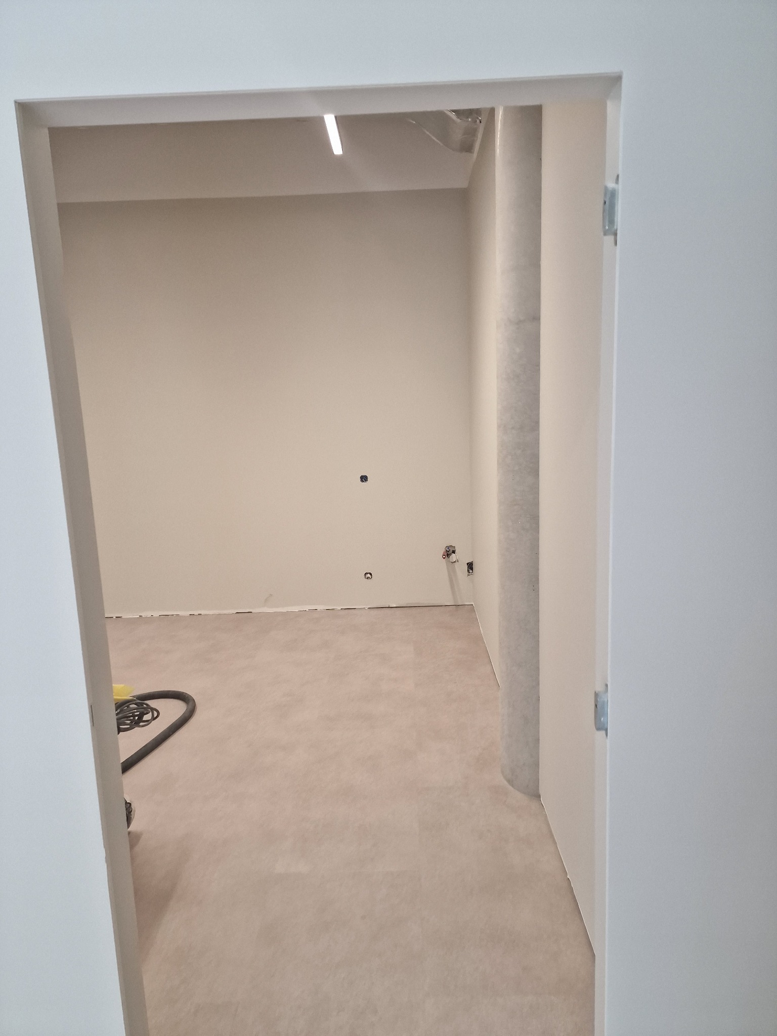 Wnętrze po remoncie: świeżo pomalowane ściany w odcieniach beżu, jasna podłoga i widoczny fragment odkurzacza. Widok zza otwartych, białych drzwi.