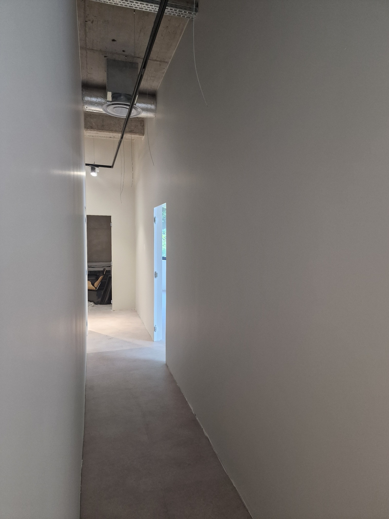 Pomalowany na gładko korytarz w neutralnym odcieniu szarości, z widocznymi elementami instalacji pod sufitem i otwartymi drzwiami w głębi, ukazującymi wnętrze.