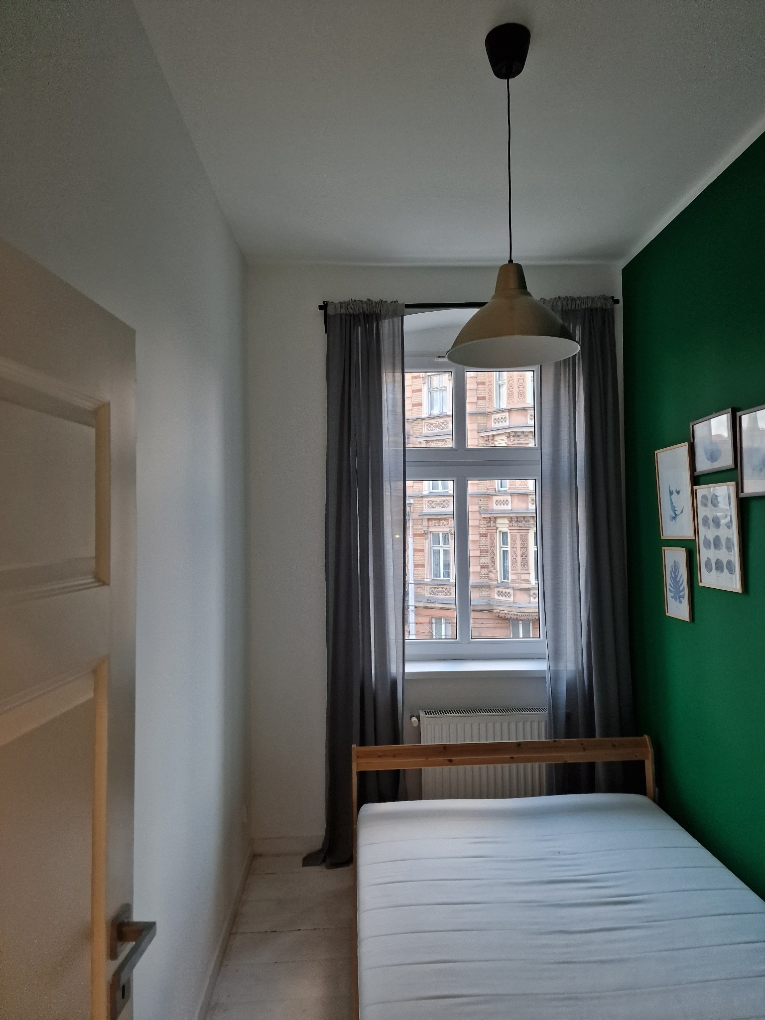 Pokój po malowaniu: zielona ściana z dekoracjami, białe ściany, okno z szarymi zasłonami, drewniane łóżko i lampa wisząca. Widok na budynek z zewnątrz.