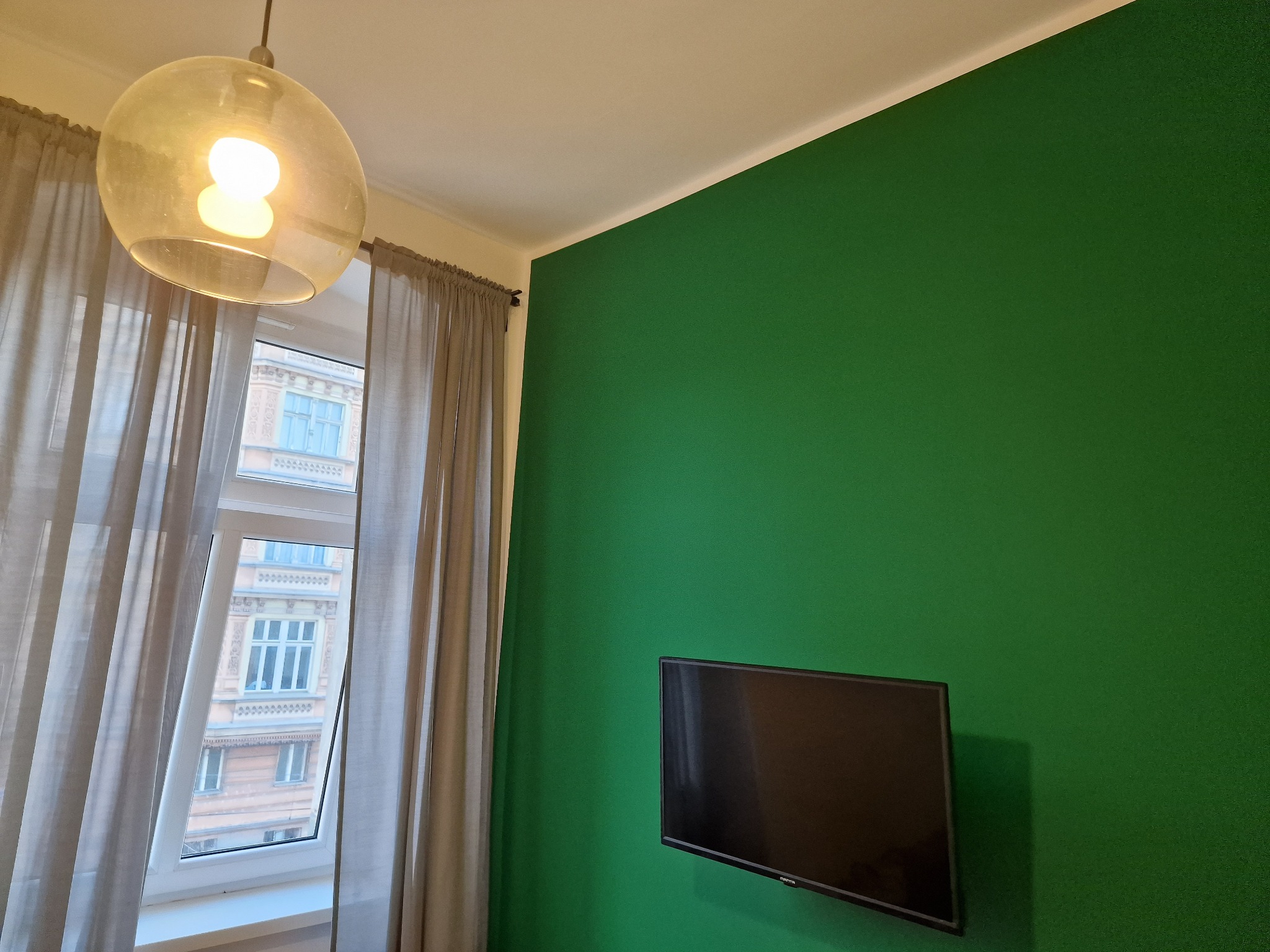 Pokój z zieloną ścianą, na której wisi telewizor. Okno z zasłonami i lampa wisząca. Widok na budynek z okna. Świeżo pomalowane ściany w pokoju.