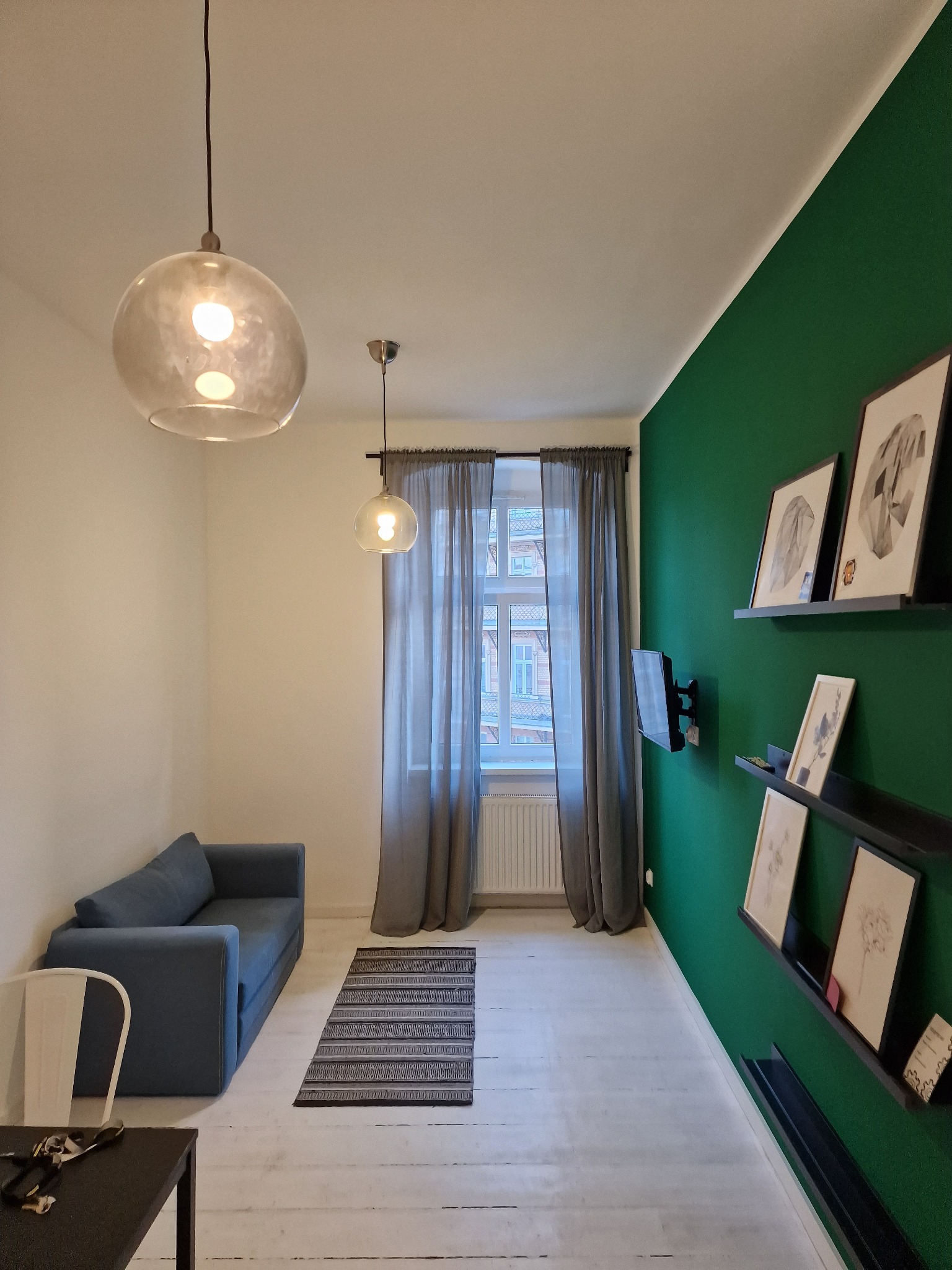 Nowoczesny salon z zieloną ścianą, szarą sofą i drewnianą podłogą. Na ścianie wiszą półki z obrazami, a okno zasłaniają szare zasłony. Dwie lampy wiszące oświetlają wnętrze.