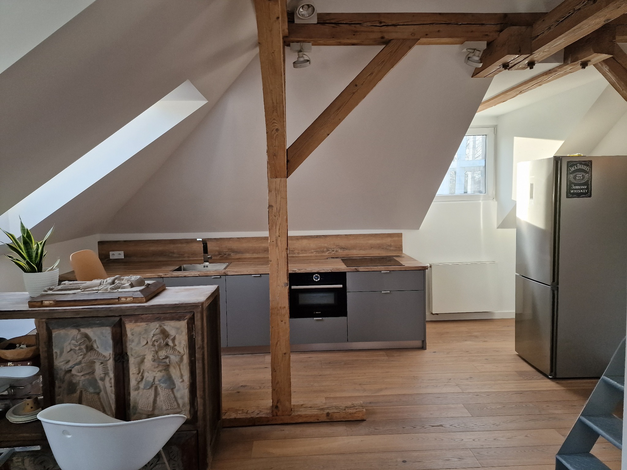Kuchnia na poddaszu z drewnianą podłogą i belkami stropowymi. Szare szafki kuchenne, sprzęt AGD i dekoracyjny kredens z rzeźbieniami.