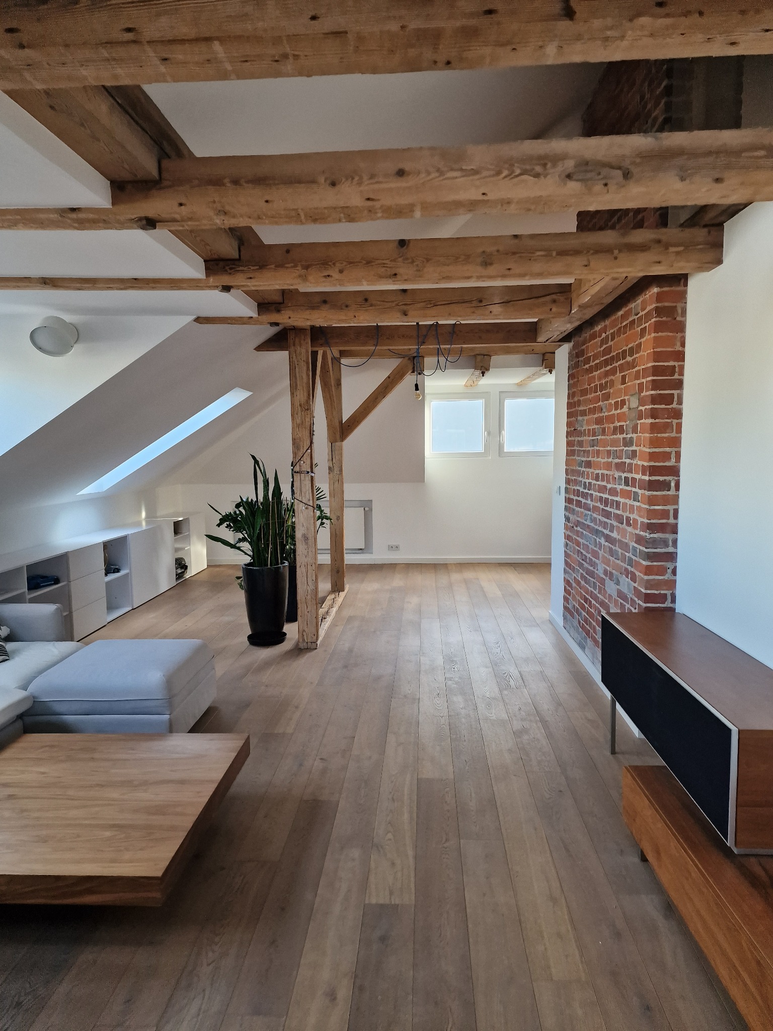 Jasne poddasze z drewnianą podłogą i belkami stropowymi, ceglana ściana, białe ściany, nowoczesne meble. Widok na przestronne, odnowione wnętrze.