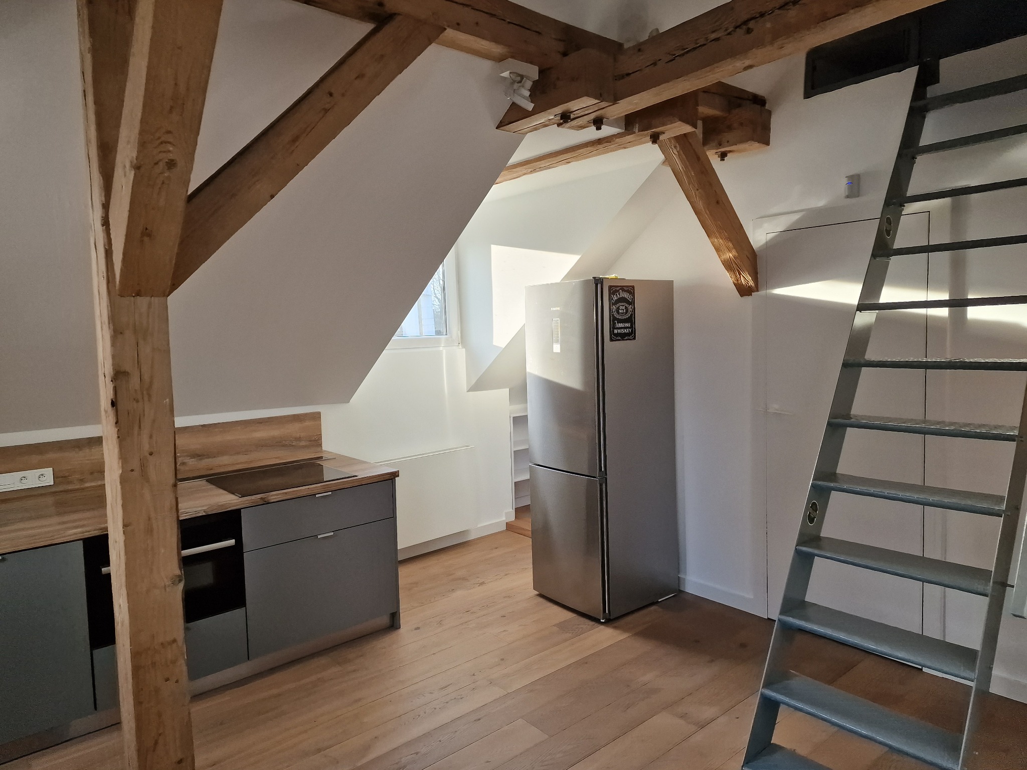 Jasne poddasze z drewnianymi belkami, szarą zabudową kuchenną, metalowymi schodami i srebrną lodówką z magnesem Jack Daniels. Parkiet z naturalnego drewna.