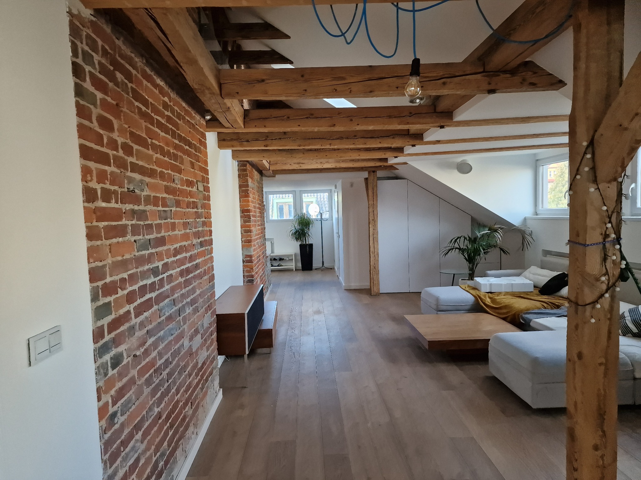 Przestronne wnętrze z ceglaną ścianą, drewnianymi belkami stropowymi i podłogą. Widoczne meble wypoczynkowe i dekoracje roślinne. Oświetlenie punktowe.