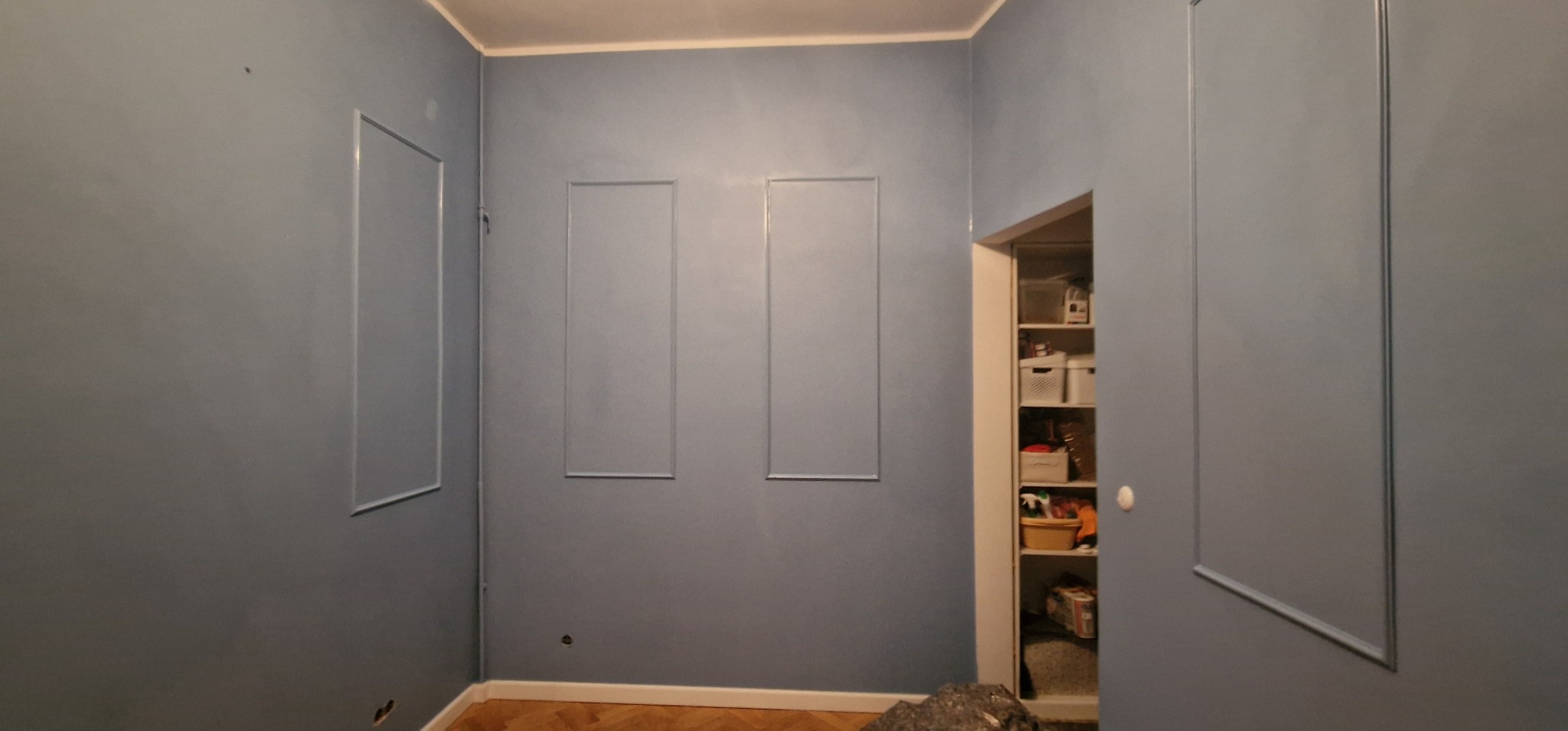 Pokój po malowaniu na szaro-niebieski kolor z dekoracyjnymi ramkami na ścianach i drewnianą podłogą. Widoczna otwarta szafa wnękowa.