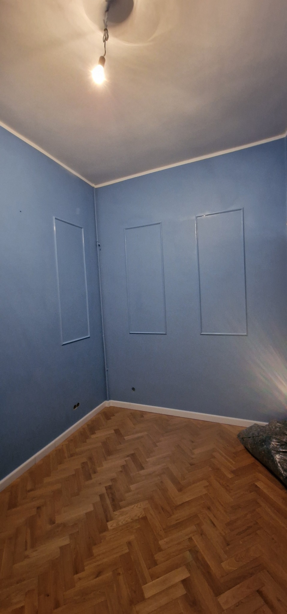 Błękitne ściany z dekoracyjnymi ramkami i parkietem w jodełkę. Widoczna żarówka. Wykończone wnętrze po malowaniu, gotowe do umeblowania.