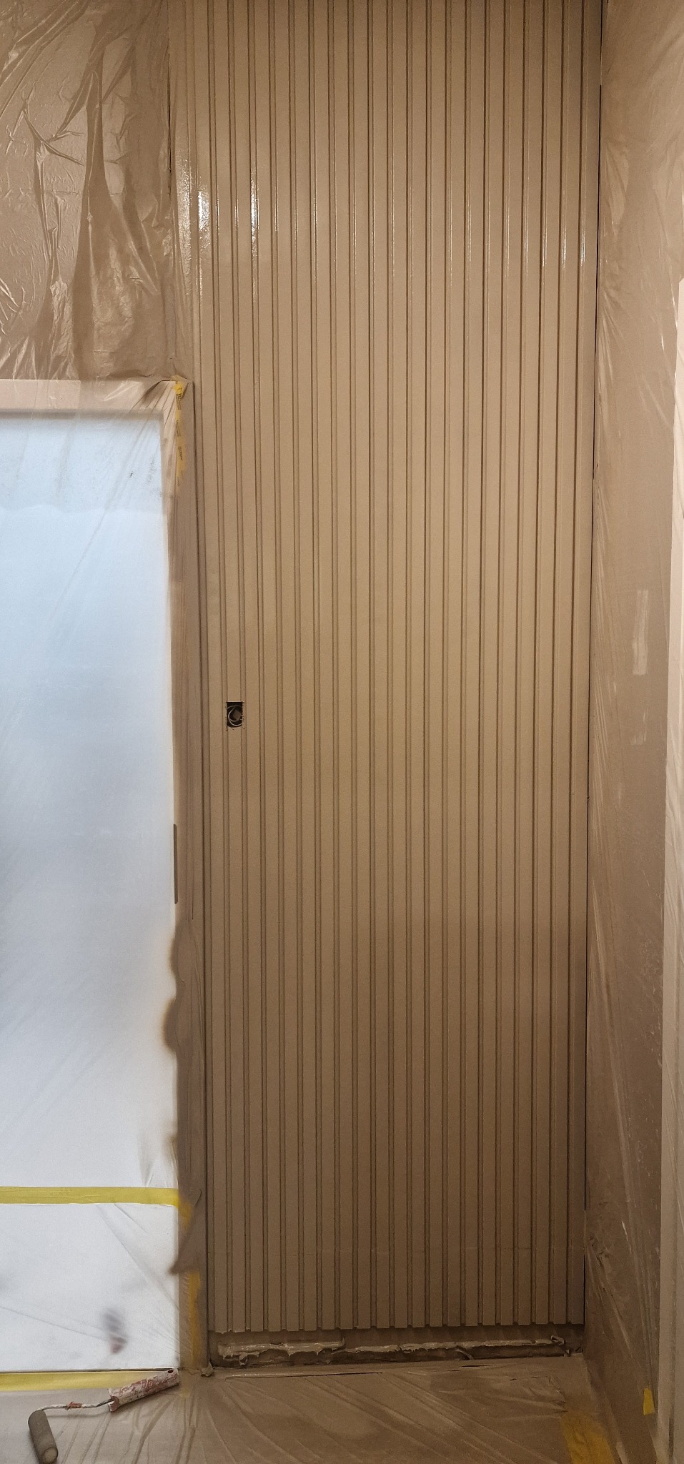 Świeżo pomalowana, beżowa ściana z pionowymi panelami, zabezpieczona folią malarską. Widoczny fragment drzwi z folią i wałek malarski na podłodze.