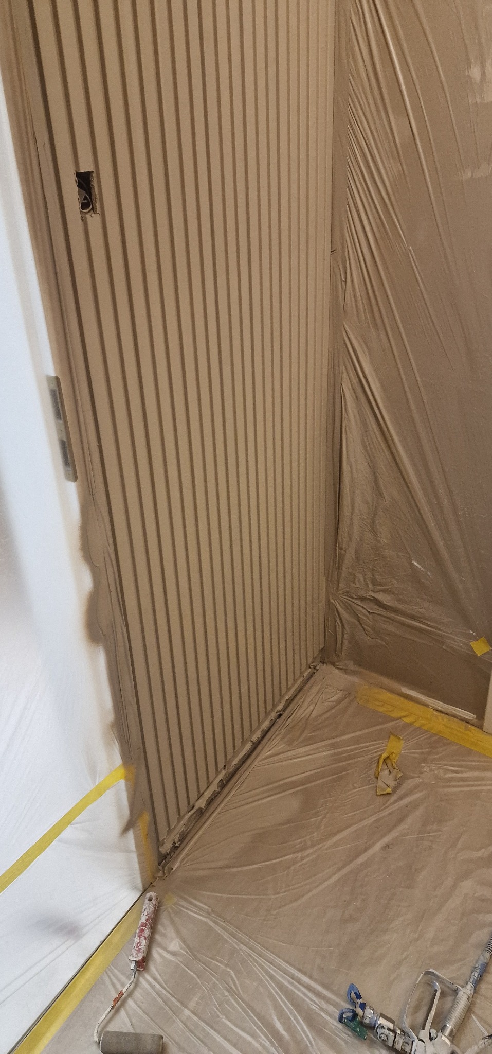 Pokój przygotowany do malowania: ściany z pionowymi żłobieniami zabezpieczone folią i taśmą malarską, widoczny wałek i pistolet natryskowy.