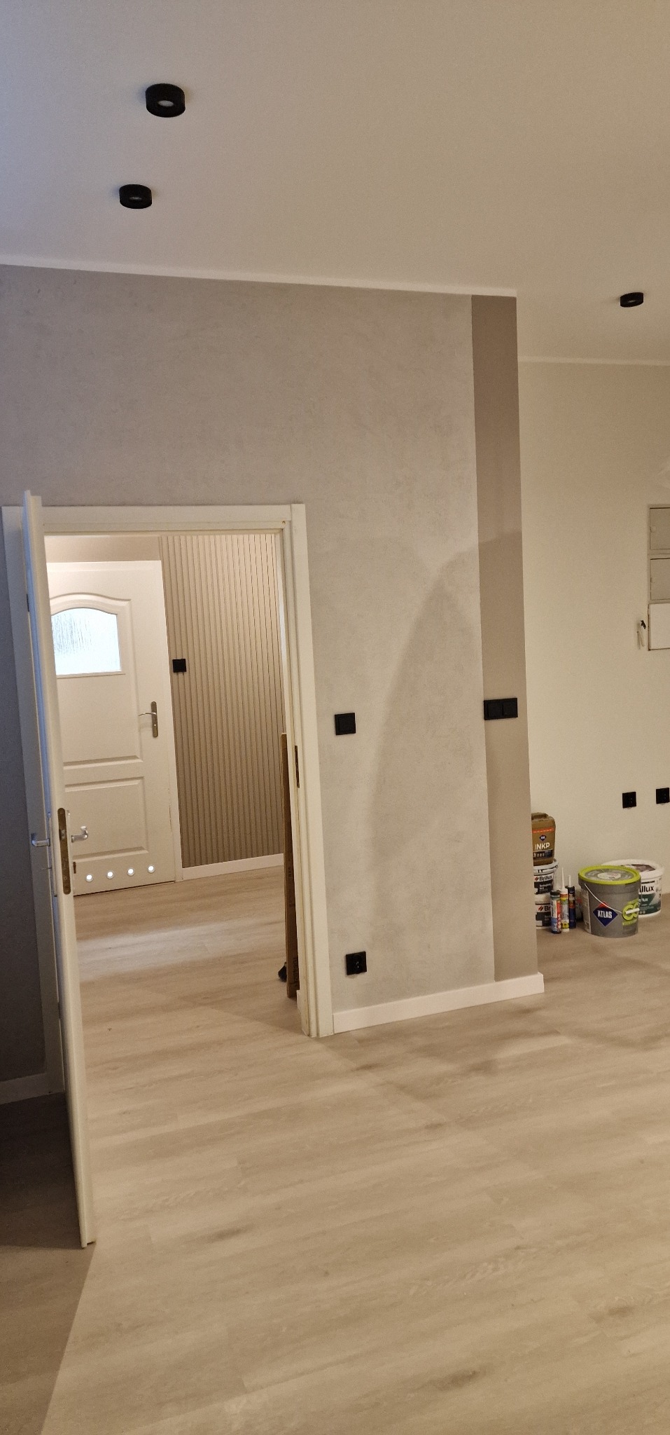 Jasne wnętrze po odświeżeniu: ściany w odcieniach szarości z akcentem beżu, widoczne puszki z farbą, podłoga z paneli. W tle otwarte drzwi do kolejnych pomieszczeń.