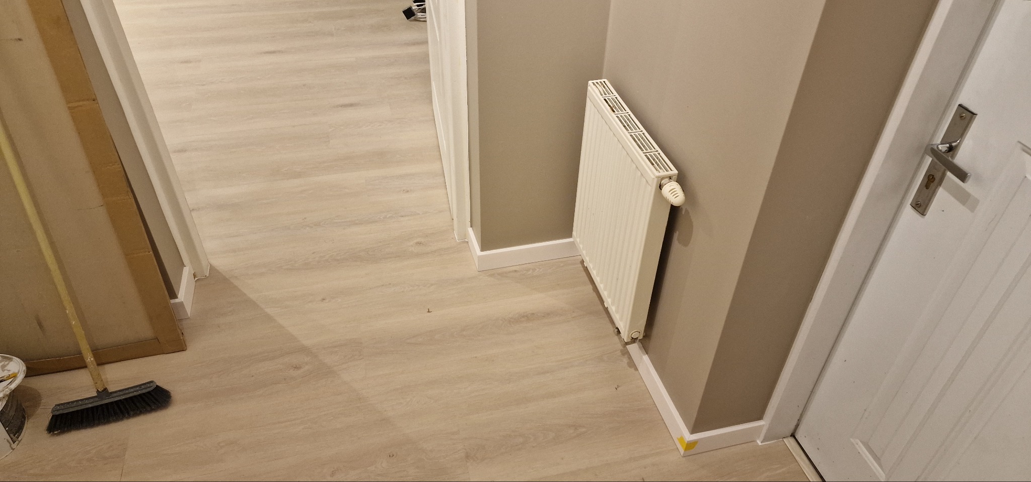 Jasne panele podłogowe po renowacji w Zamościu, widok z góry. Ściany pomalowane na szaro, białe listwy przypodłogowe i grzejnik. Częściowo widoczna biała stolarka drzwiowa.