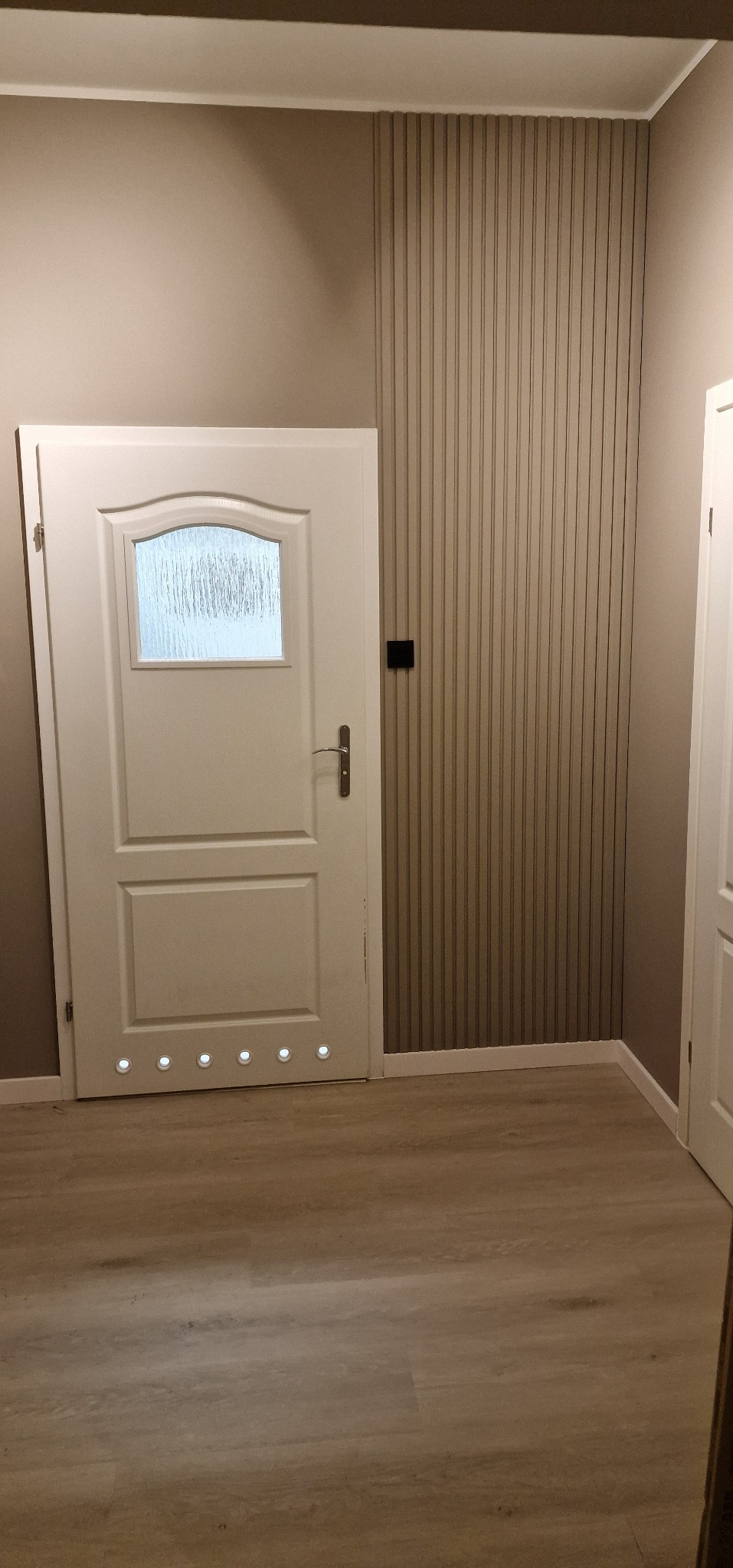 Białe drzwi z szybą i podświetleniem LED u dołu, zamontowane w odnowionym wnętrzu z panelami ściennymi i drewnianą podłogą. Widok na fragment drugich drzwi.