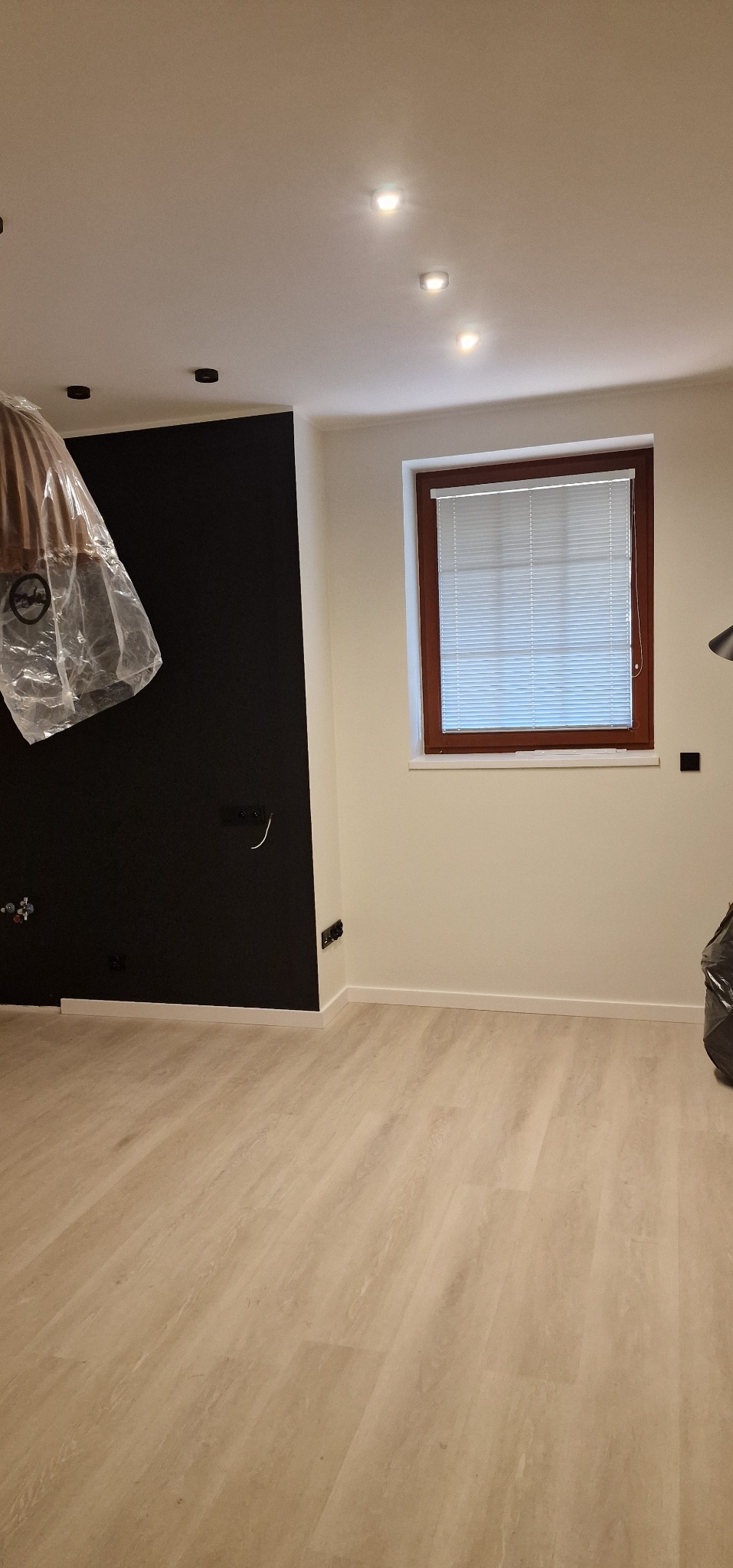 Pokój po remoncie: jasne panele podłogowe, czarna ściana z gniazdkami, okno z żaluzjami. Część lampy wiszącej zabezpieczona folią. Widok na odnowione wnętrze.