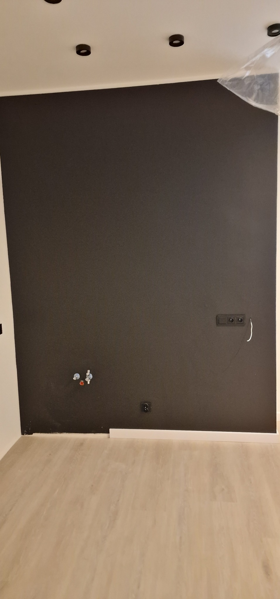 Ciemna ściana po malowaniu z widocznymi gniazdkami, rurami i listwą przypodłogową. Na podłodze jasne panele. Fragment folii ochronnej na górze ściany.