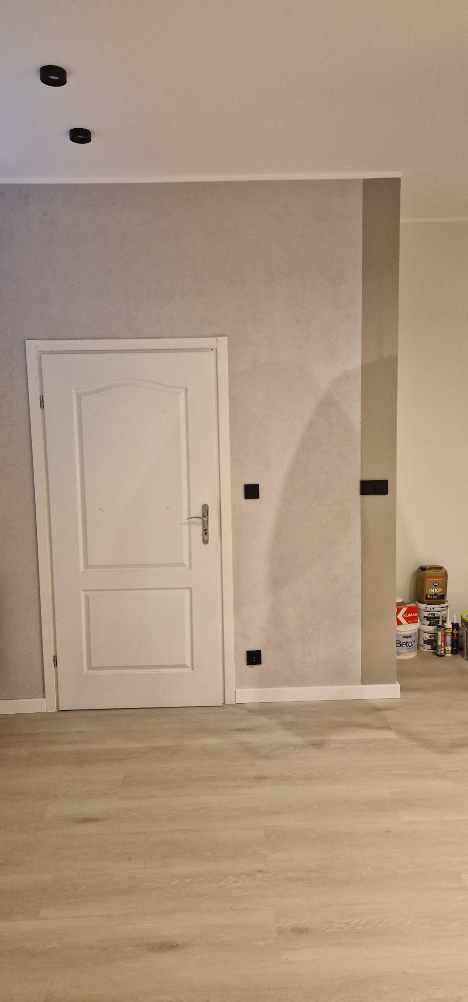 Wykończone wnętrze: białe drzwi, ściana z efektem betonu w odcieniach szarości, panele podłogowe. Widoczne puszki z farbami i narzędzia malarskie przy ścianie.