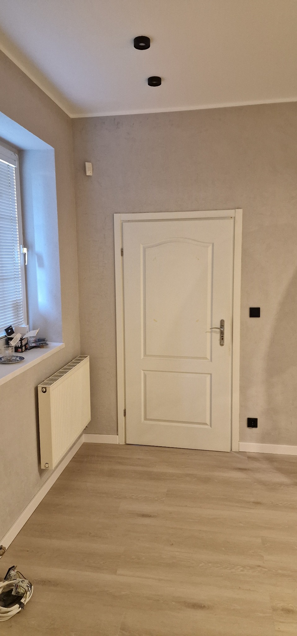 Jasne wnętrze po remoncie: białe drzwi, grzejnik pod oknem z żaluzjami, panele podłogowe i szare ściany z widocznymi gniazdkami. Dwa czarne punkty oświetleniowe na suficie.