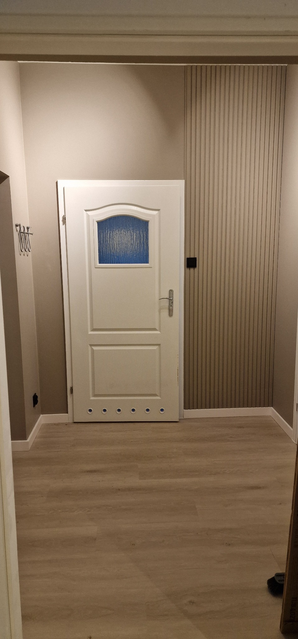 Białe drzwi z szybą w odcieniach błękitu, zamontowane w korytarzu z panelami podłogowymi i dekoracyjną ścianą z pionowych listew w kolorze beżowym.