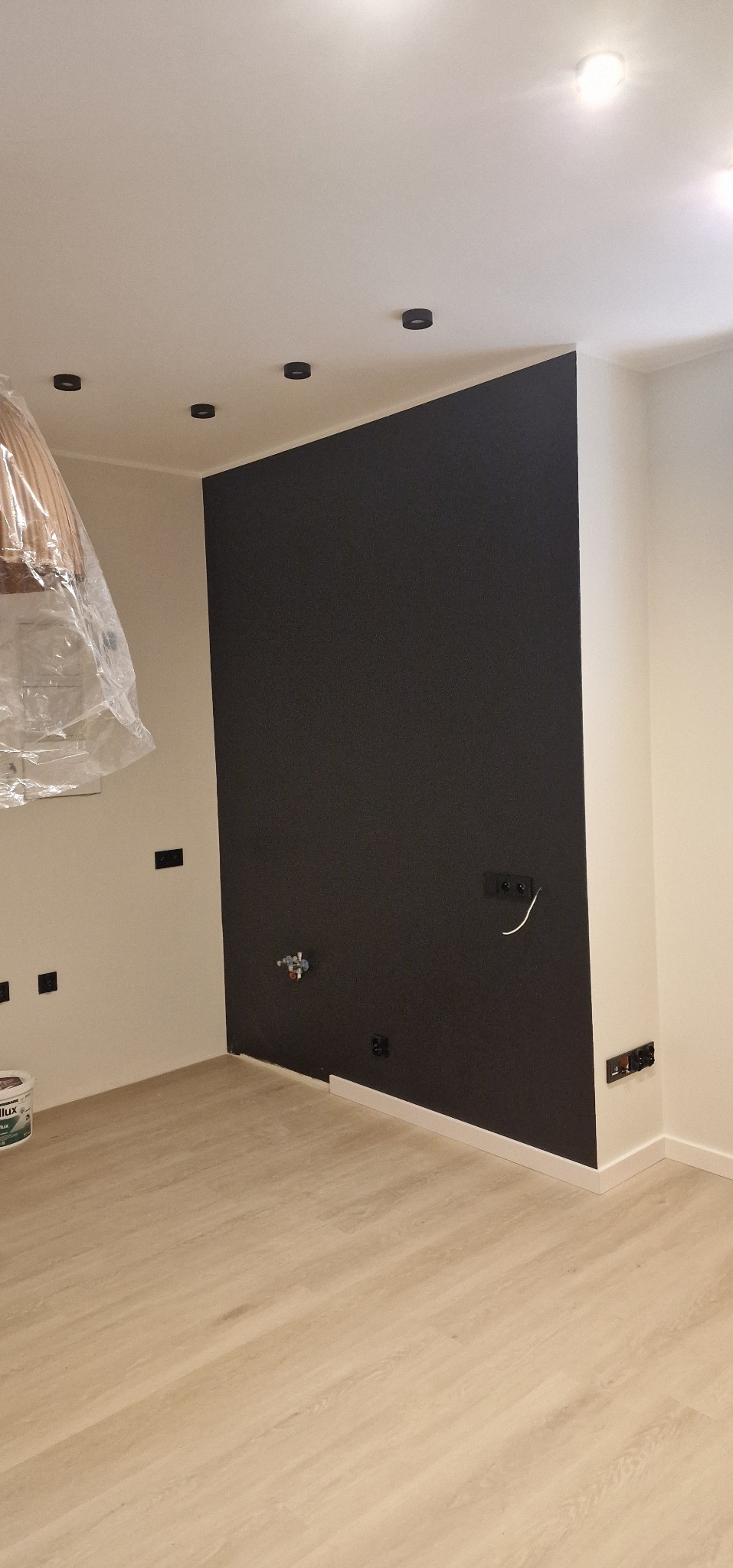 Pokój po remoncie: czarna ściana kontrastuje z jasnymi panelami podłogowymi i białymi ścianami. Widoczne gniazdka elektryczne i lampy sufitowe. Część lampy zakryta folią.