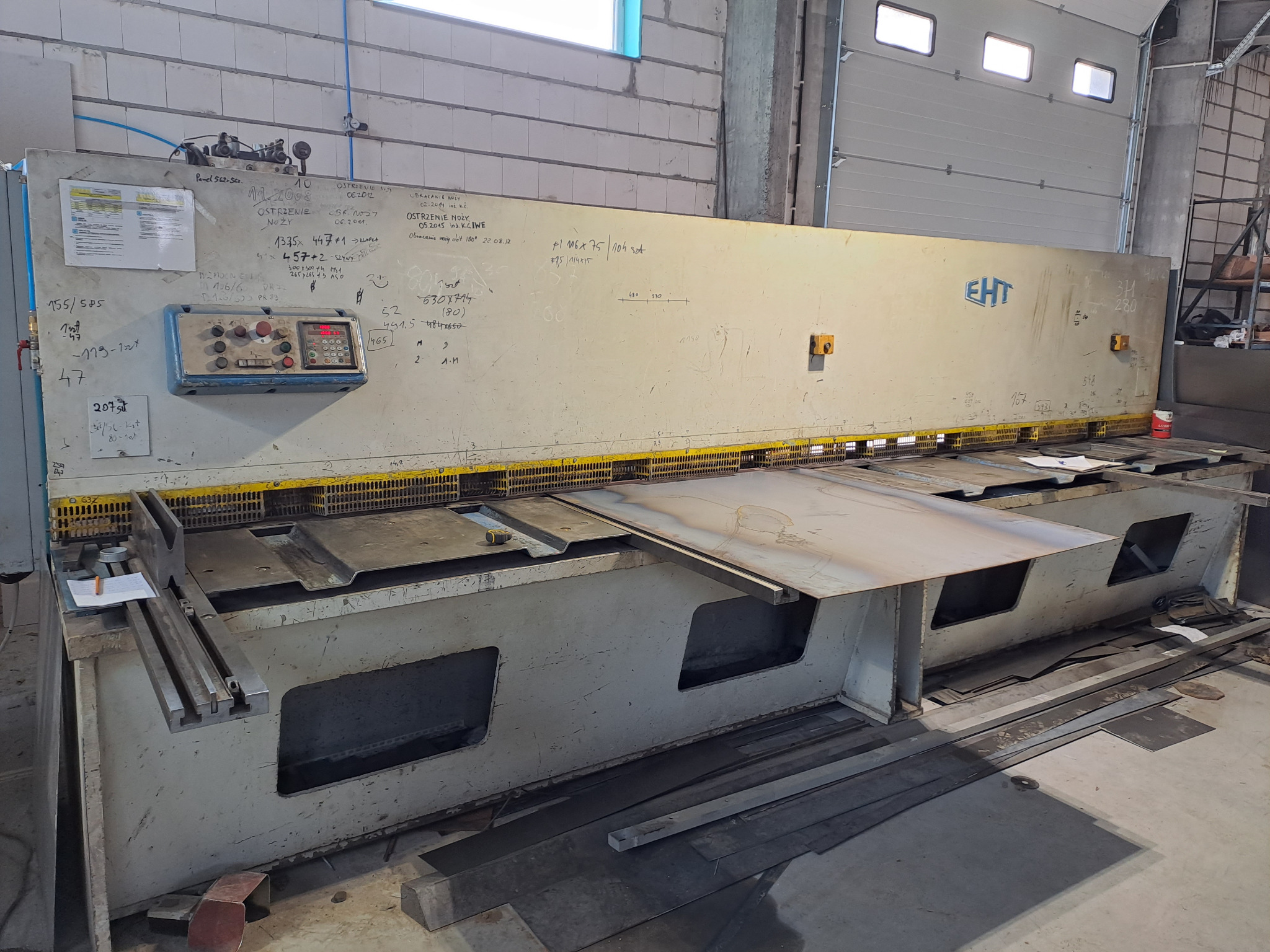 Duża, przemysłowa gilotyna do cięcia metalu, z widocznymi śladami użytkowania i notatkami technicznymi na obudowie, w hali produkcyjnej.