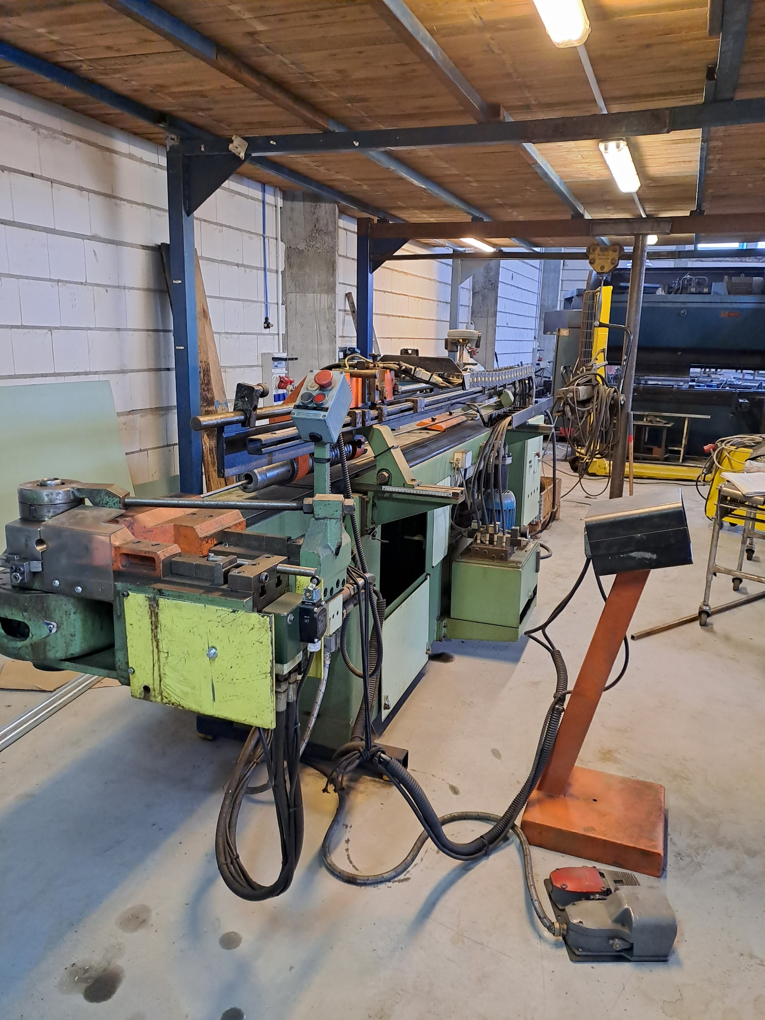 Duża, zielono-żółta giętarka do metalu w warsztacie, z widocznymi przewodami hydraulicznymi i panelem sterowania, pod drewnianym stropem z oświetleniem.