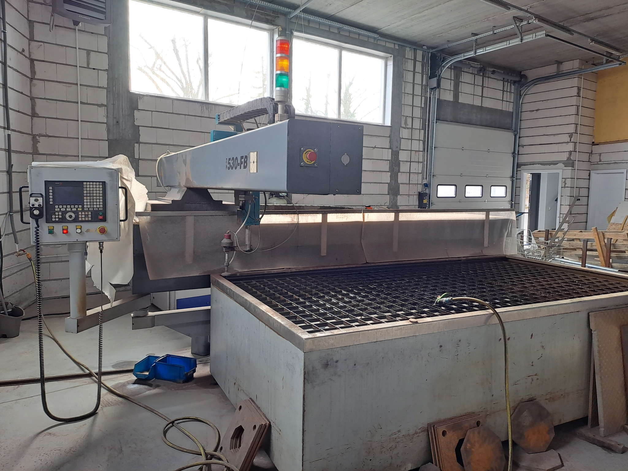 Wycinarka wodna CNC model 1530-FB w hali produkcyjnej, z widocznym pulpitem sterowniczym, stołem roboczym z rusztem i sygnalizacją świetlną. Na podłodze leżą wycięte elementy stalowe.