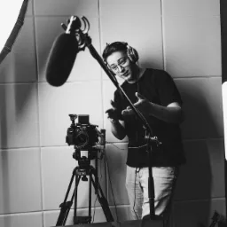 Czarno-białe zdjęcie mężczyzny w okularach i słuchawkach gestykulującego przed kamerą z mikrofonem w studio z panelami akustycznymi i oświetleniem.