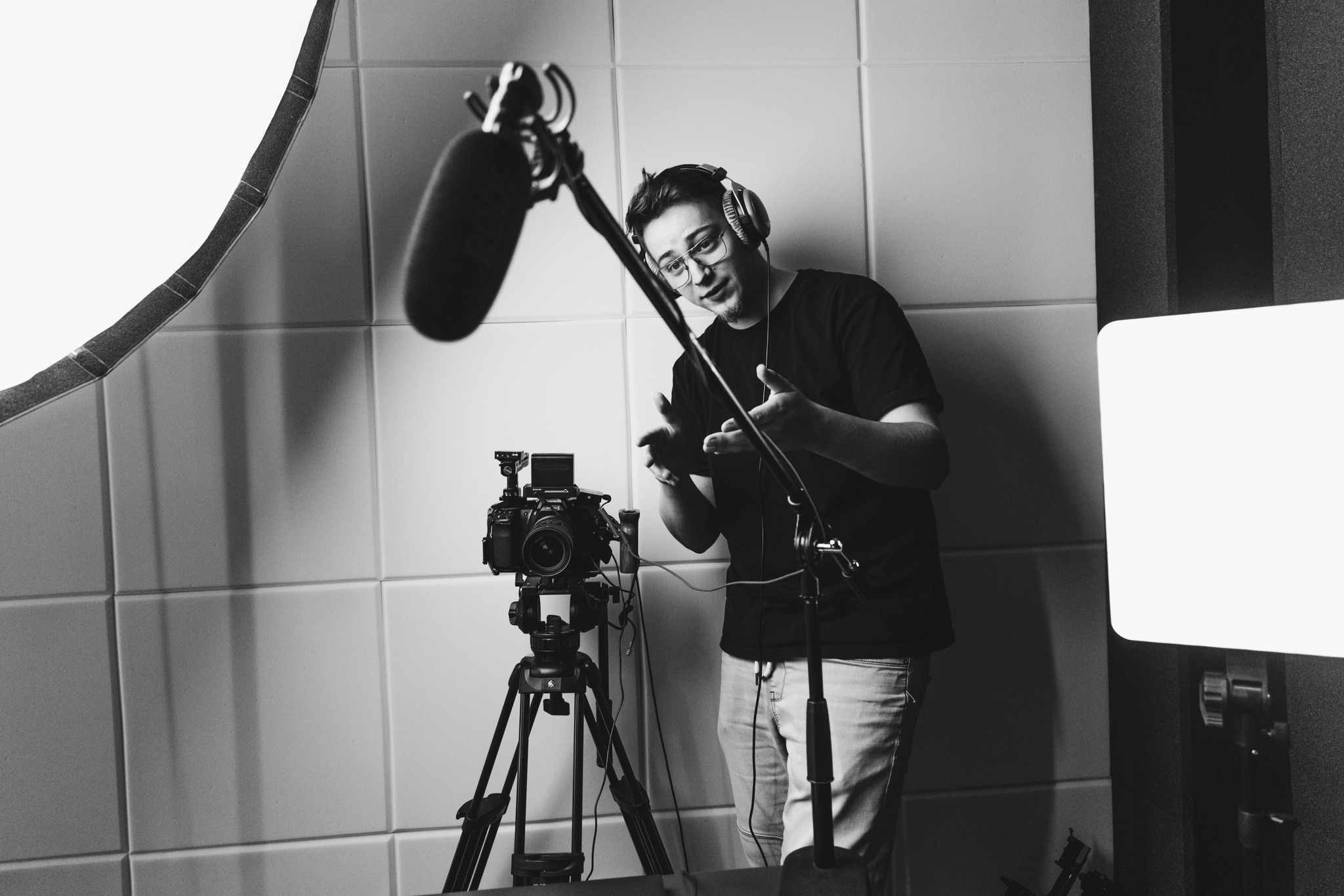 Czarno-białe zdjęcie mężczyzny w okularach i słuchawkach gestykulującego przed kamerą z mikrofonem w studio z panelami akustycznymi i oświetleniem.