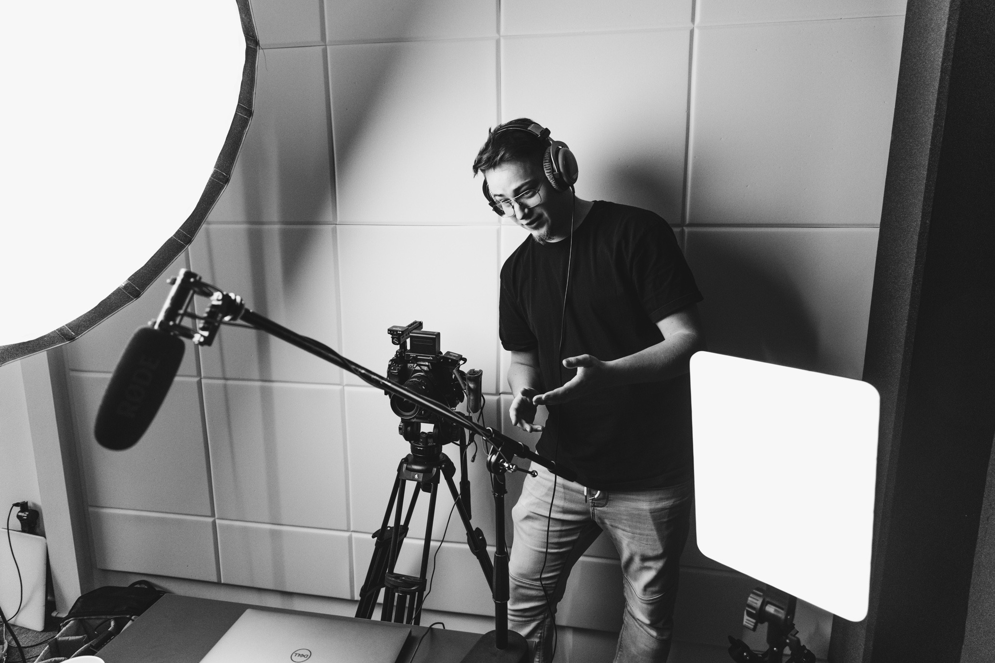 Czarno-białe zdjęcie mężczyzny w słuchawkach, stojącego w studio z kamerą na statywie, mikrofonem Rode i lampami LED, przygotowującego się do nagrania.