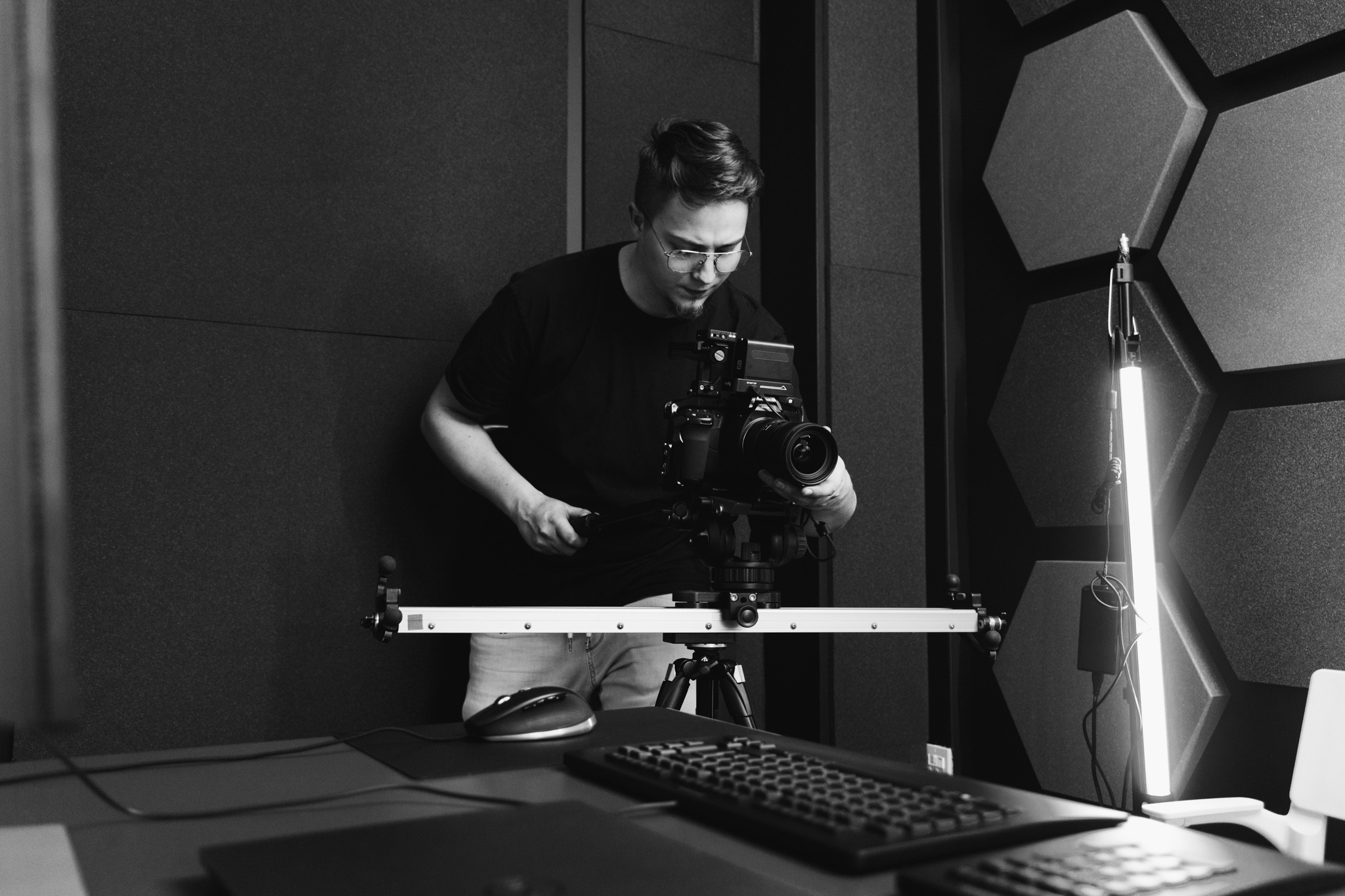 Czarno-białe zdjęcie filmowca z okularami, regulującego kamerę na statywie w studio z panelami akustycznymi w kształcie plastrów miodu, oświetlonego listwą LED.