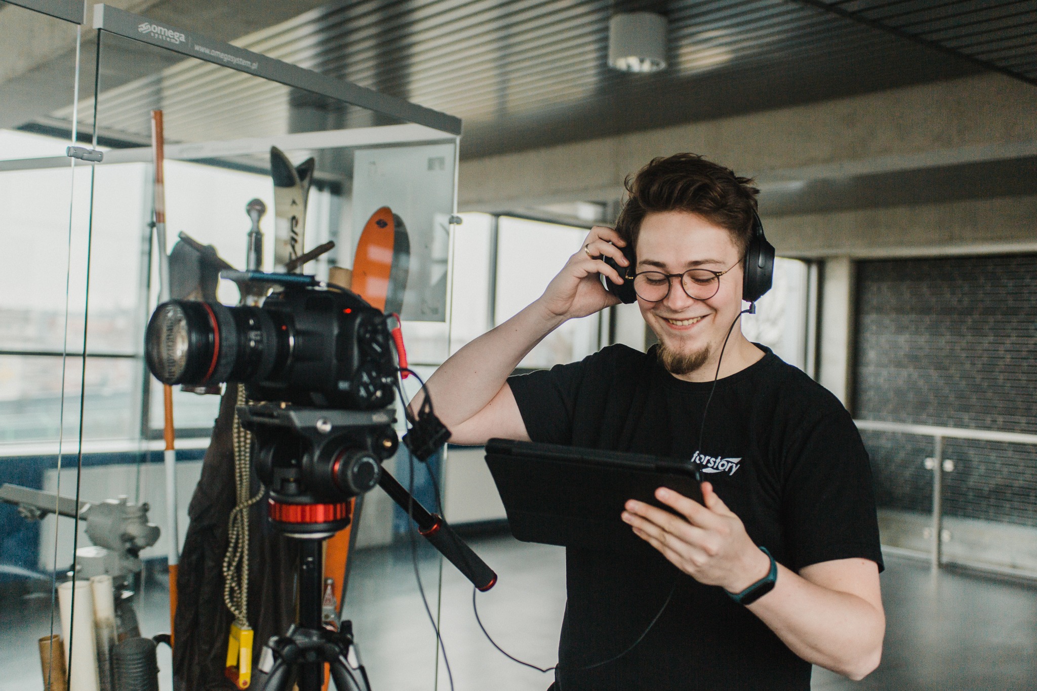 Uśmiechnięty mężczyzna w słuchawkach, z tabletem w ręku, stoi obok kamery na statywie; w tle rekwizyty i przeszklona ściana.