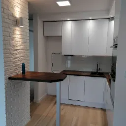 Nowoczesna kuchnia z białymi szafkami, drewnianym blatem i ścianą z białej cegły, na której umieszczona jest półka barowa z turkusową butelką.