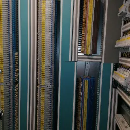 Szczegółowe ujęcie rozdzielnicy elektrycznej z rzędami zacisków, każdy oznaczony żółtą etykietą, umieszczonych wewnątrz metalowej szafy z turkusowymi panelami.