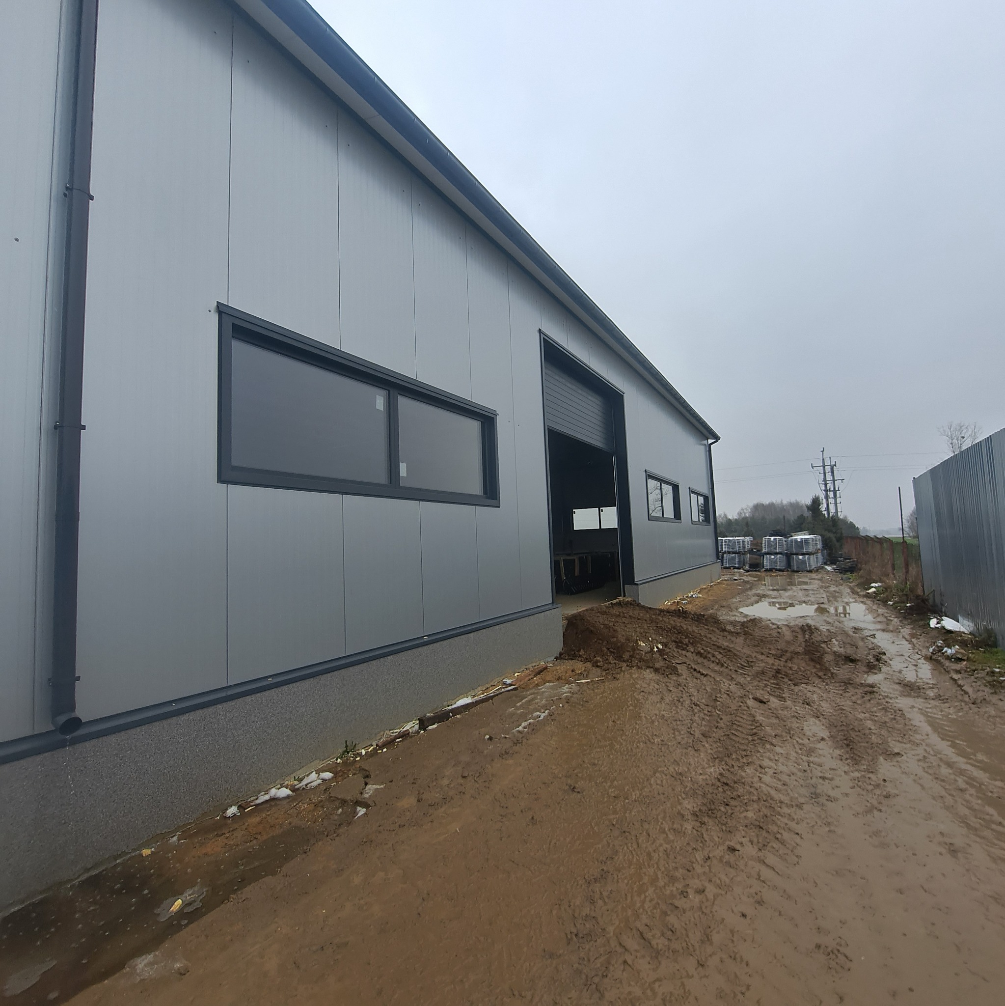 Nowoczesny budynek przemysłowy z szarych płyt warstwowych, z otwartą bramą rolowaną i stertą materiałów budowlanych na zewnątrz, widoczne błotniste podłoże.